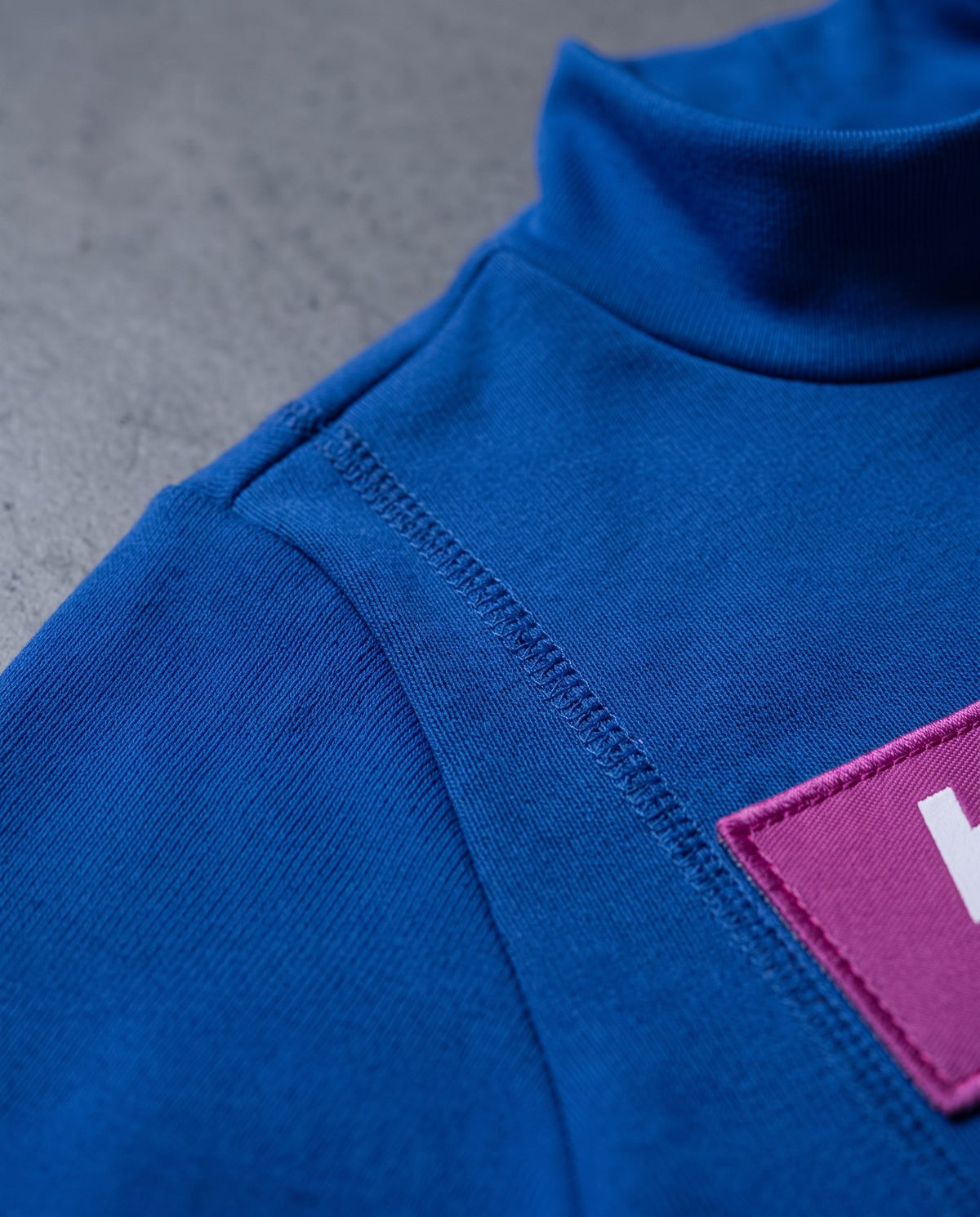 Materialnahaufnahme: Rollkragenkante, Flachnaht und Sponsorenfeld des blauen Bodys
